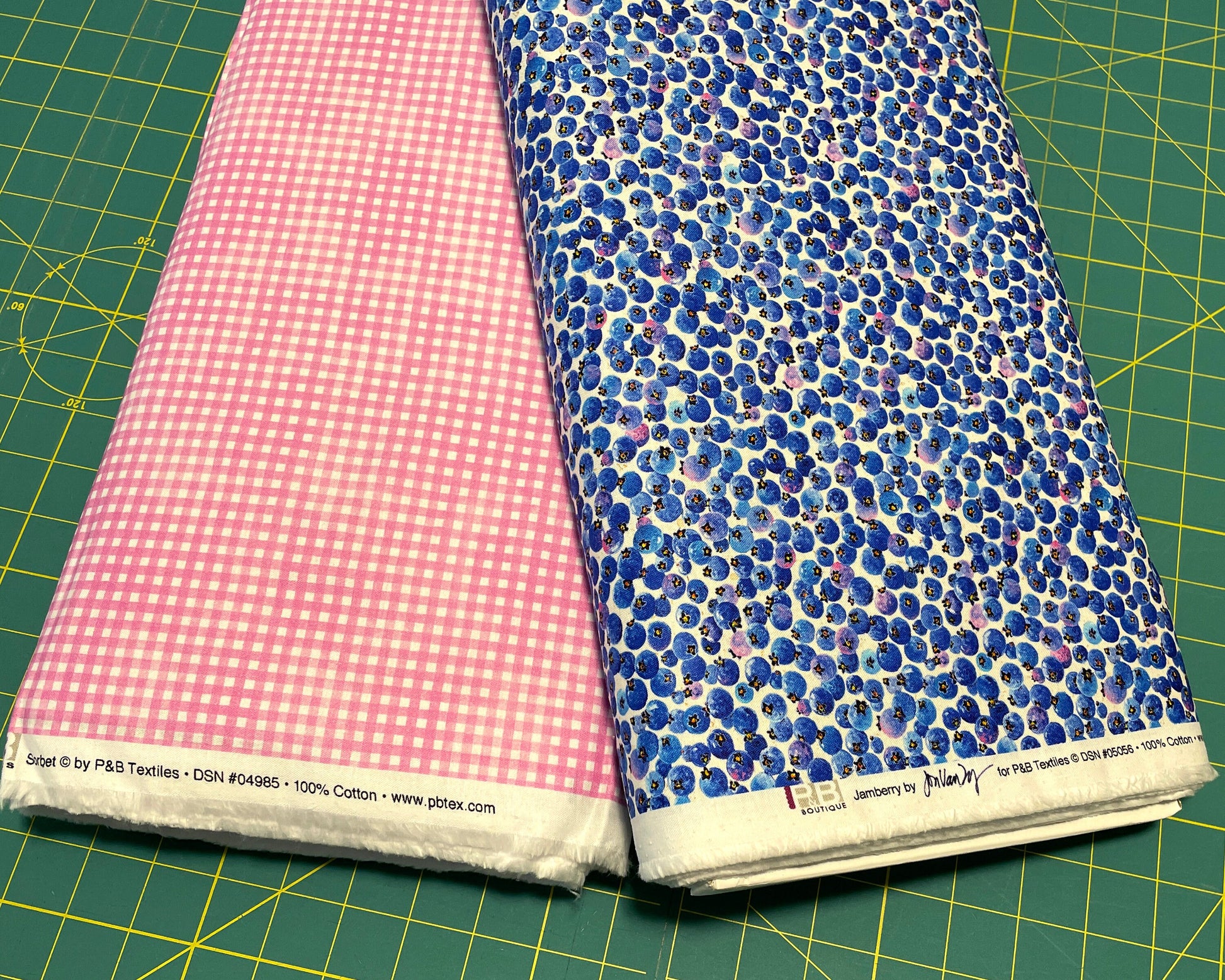 Two bolts of fabric including one with Tossed blueberries on a white background next to a Fuchsia Gingham coordinating fabric